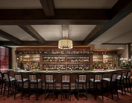 A cozy bar with a long wooden counter, many bottles on shelves, stools, warm lighting, and a hanging chandelier.