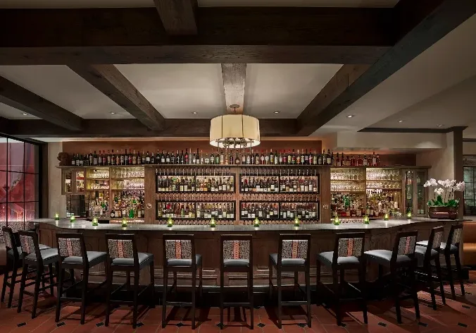 A cozy bar interior packed with colorful liquor bottles behind a wooden counter and lined with high stools.
