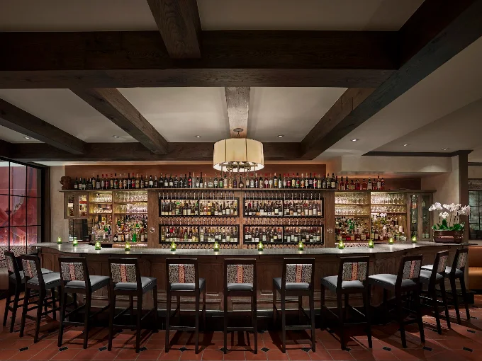 A cozy bar interior packed with colorful liquor bottles behind a wooden counter and lined with high stools.