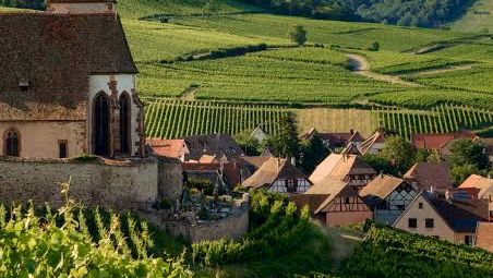 A hilltop village with vineyards and a church featuring a tall red-brown steeple, surrounded by green forested mountains under a blue sky.