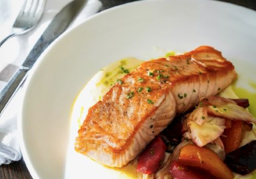 A plated seared salmon with roasted vegetables on a bed of greens, with a wine glass and forks on the table.