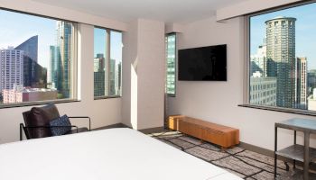 The image shows a modern hotel room with a bed, TV, and city views from large windows, featuring tall buildings and a minimalist design.