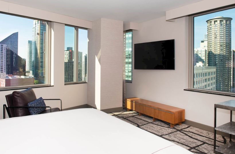 The image shows a modern hotel room with a bed, TV, and city views from large windows, featuring tall buildings and a minimalist design.