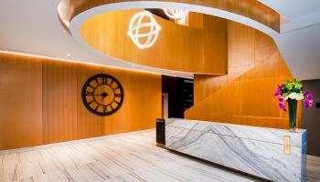 The image shows a modern lobby with a marble reception desk, wooden walls, a large clock, spiral lighting, and flowers on the desk.