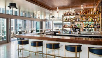 A modern bar with high stools, a selection of bottles on shelves, and large windows providing a view of the city skyline.