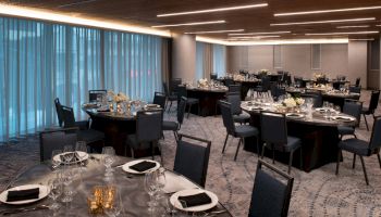 A banquet hall with elegantly set tables, featuring plates, glasses, and napkins, under ambient lighting and next to large windows.