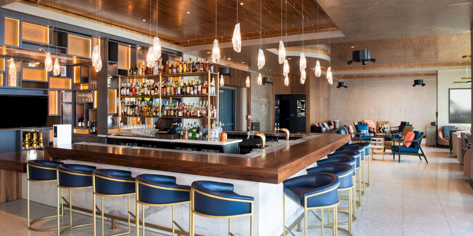 This image shows a modern bar with a wooden counter, blue stools, hanging lights, and shelves stocked with bottles.