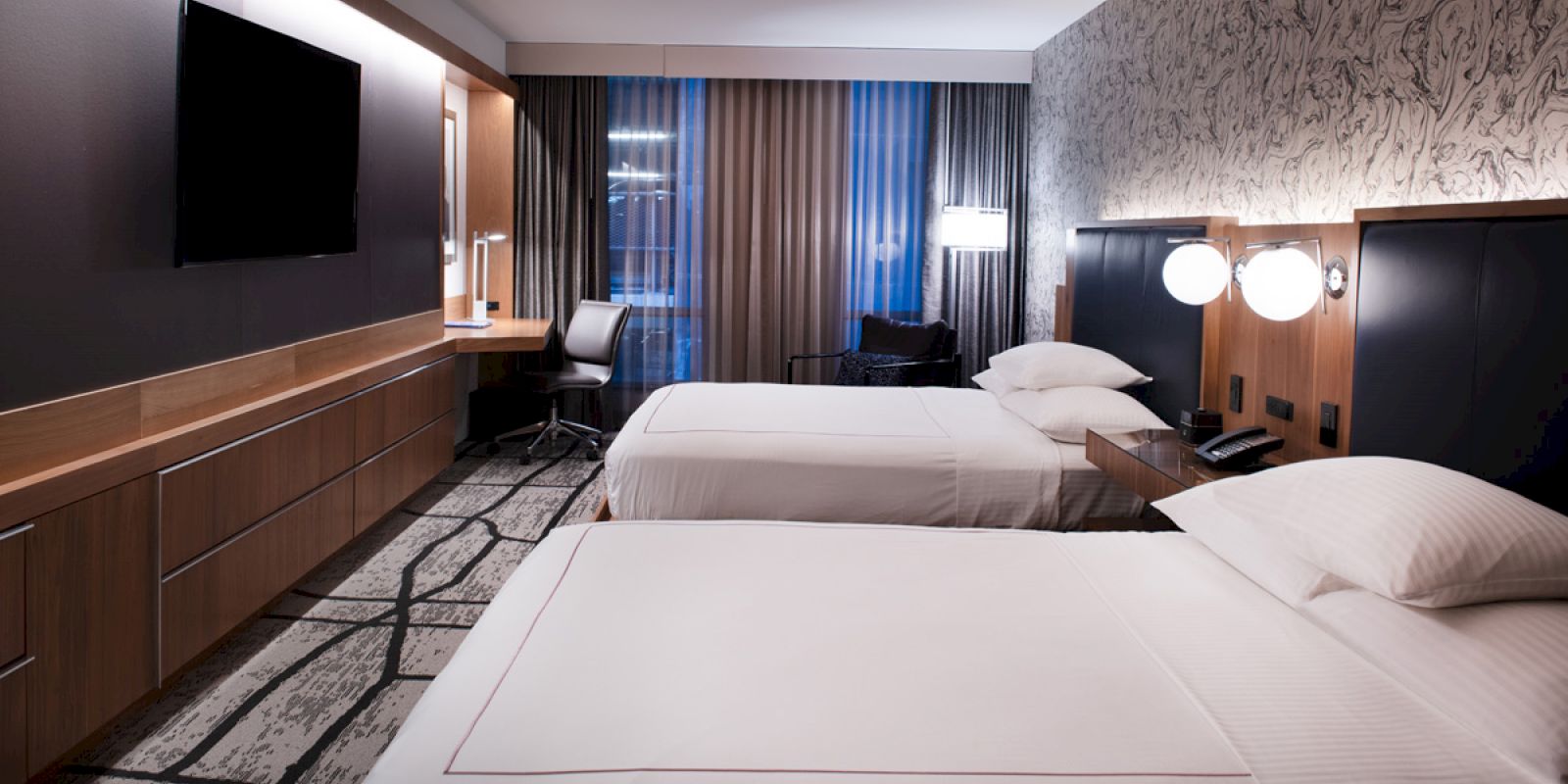 A hotel room with two beds, a TV, desk, and curtains can be seen in this image. The room has a modern design and soft lighting.