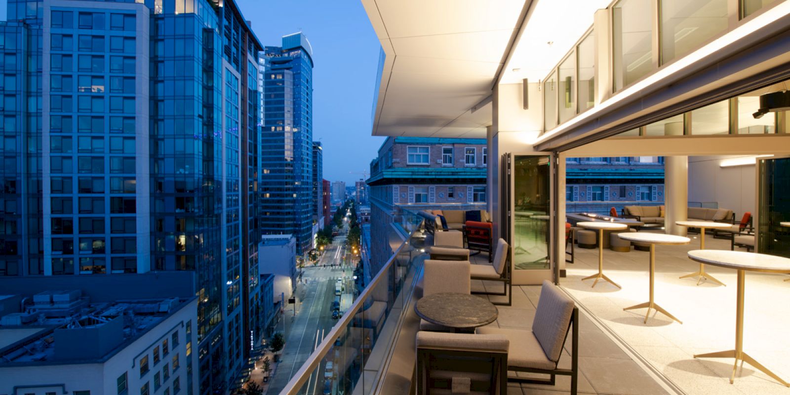 A modern cityscape at dusk featuring tall buildings, a lit outdoor terrace with seating, and a view of a street below.