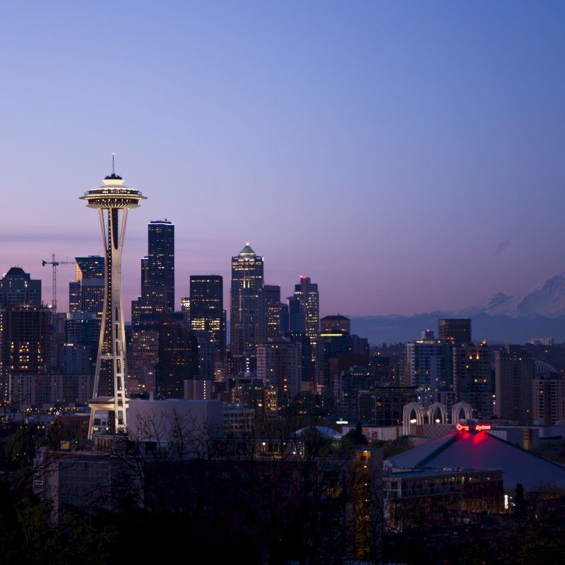 Seattle skyline at dusk with the Space Needle and Mount Rainier in the background, set against a purple-blue sky.