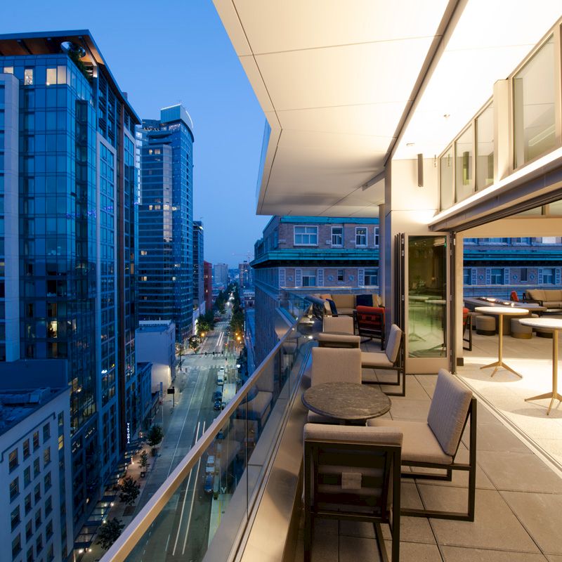 A modern rooftop terrace at dusk overlooks a city street, featuring seating and tables, with high-rise buildings in the background.