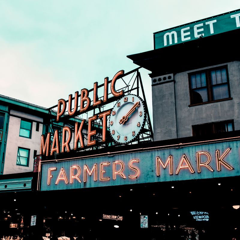 The image shows a public market sign with a clock and "Farmers Market" in neon lights, on a building with signs saying "Meet the...".