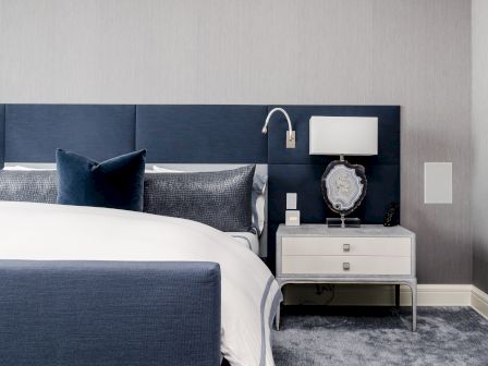A modern bedroom with a blue headboard, pillows, a white nightstand, a lamp, and a decorative mirror displayed on a gray carpet.