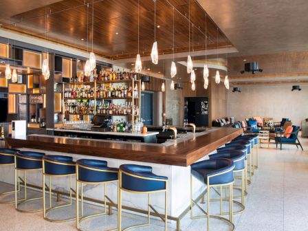 The image shows a modern bar with a large counter, blue stools, hanging lights, and shelves stocked with various bottles.