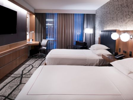 The image shows a hotel room with two beds, a flat-screen TV, a desk, and modern lighting and decor.