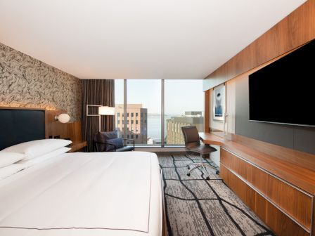 The image shows a modern hotel room with a large bed, flat-screen TV, desk, and window offering a city view.