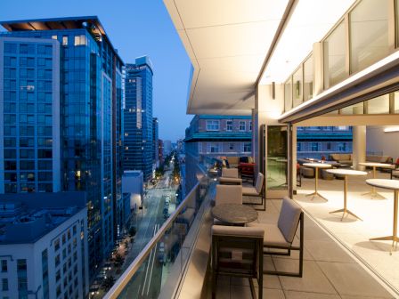 A modern balcony overlooks a cityscape with tall buildings and a street at dusk, featuring seating areas and soft lighting.