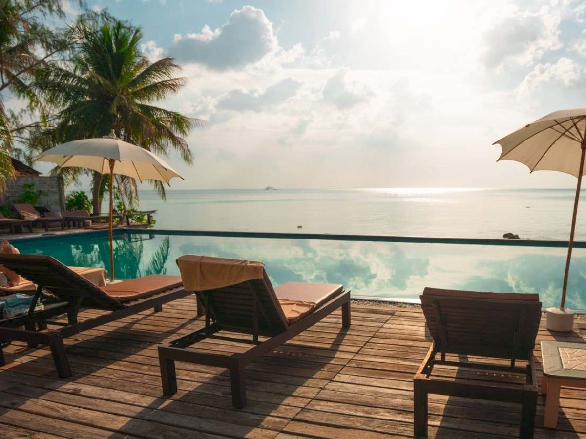Poolside scene with lounge chairs, umbrellas, and palm trees overlooking a serene water body reflecting the sky and clouds.