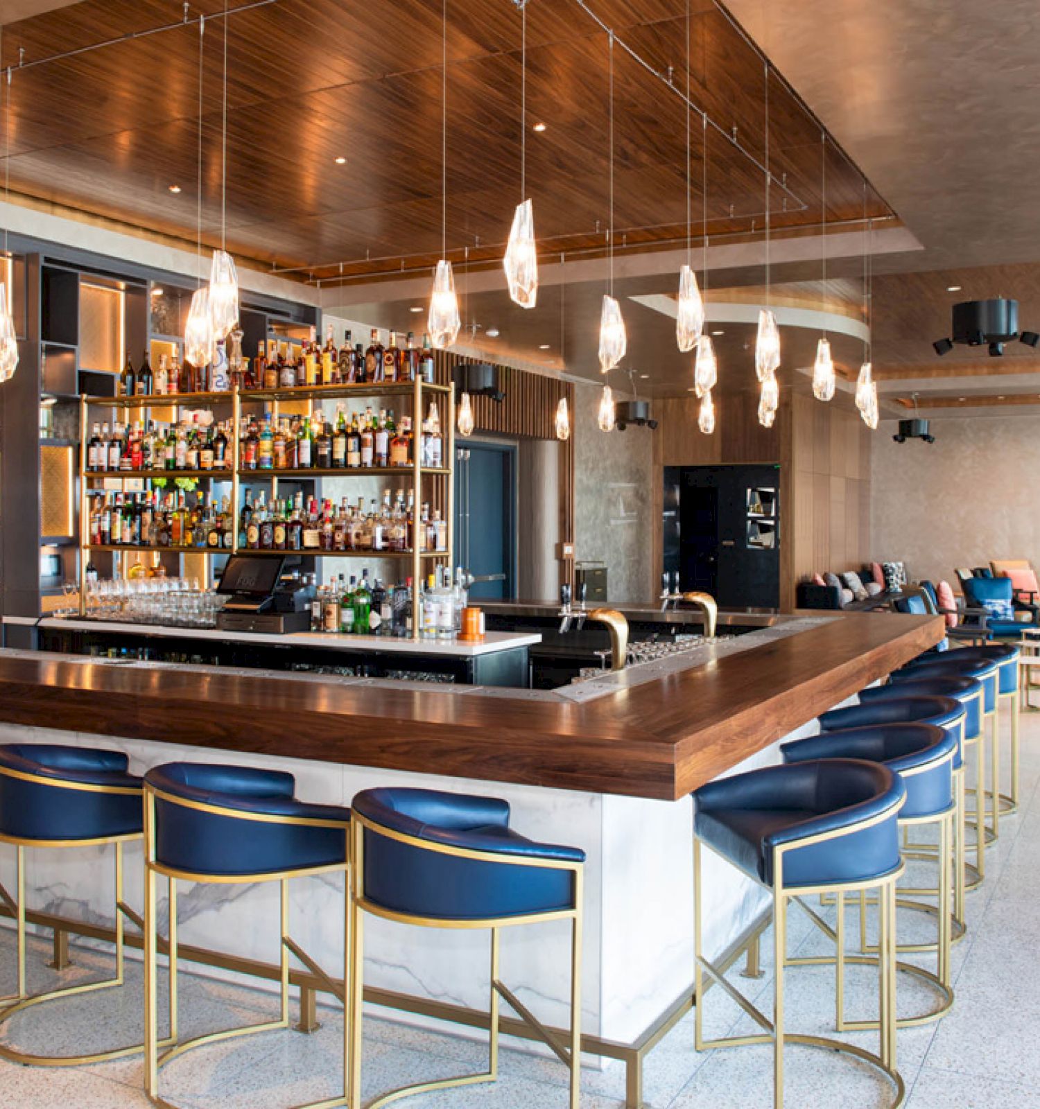 The image shows a modern bar with blue stools, overhead lighting, and a well-stocked bar counter in an open, stylish space.
