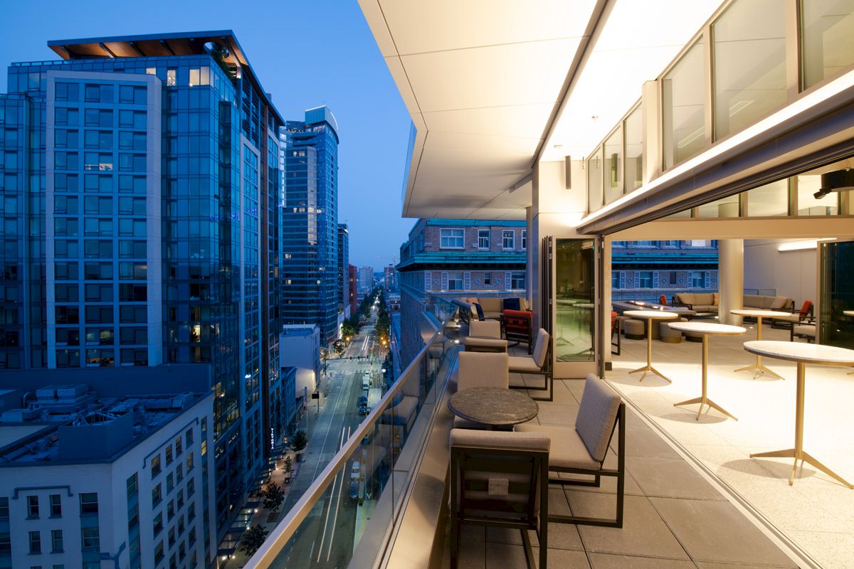 A modern rooftop terrace with seating offers a view of a cityscape with tall buildings at dusk, creating a stylish urban ambiance.