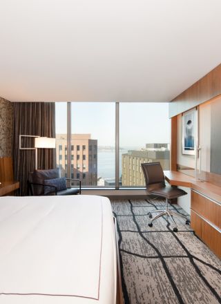 A modern hotel room features a large bed, flat-screen TV, desk, chair, and city view through a floor-to-ceiling window.