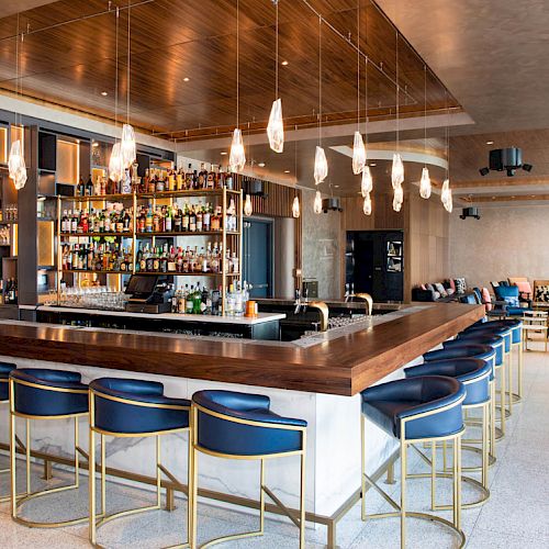 The image shows a modern bar with blue cushioned stools, a well-stocked alcohol shelf, and pendant lighting in a stylish lounge setting.