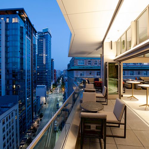 A modern cityscape at dusk featuring a high-rise balcony with seating, overlooking streets and buildings under a blue evening sky.