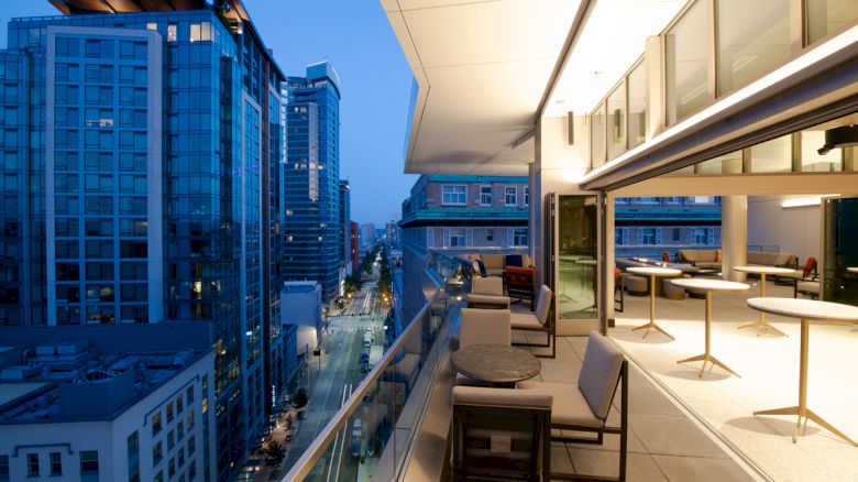 Modern city skyline view from a rooftop terrace at dusk, featuring outdoor seating and tables under ambient lighting.