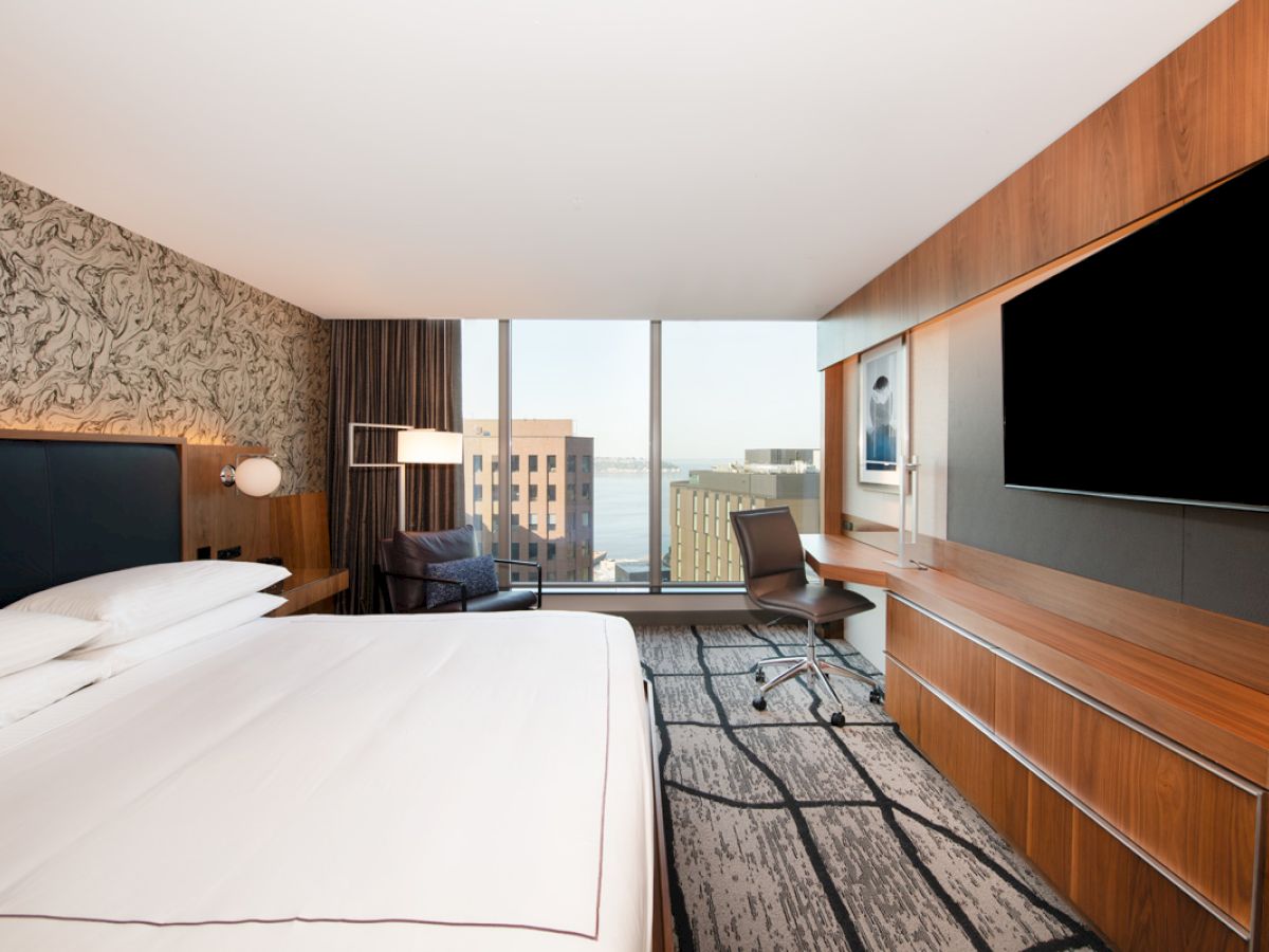 A modern hotel room with a bed, large TV, desk, chair, and a window showing a cityscape view in the background.