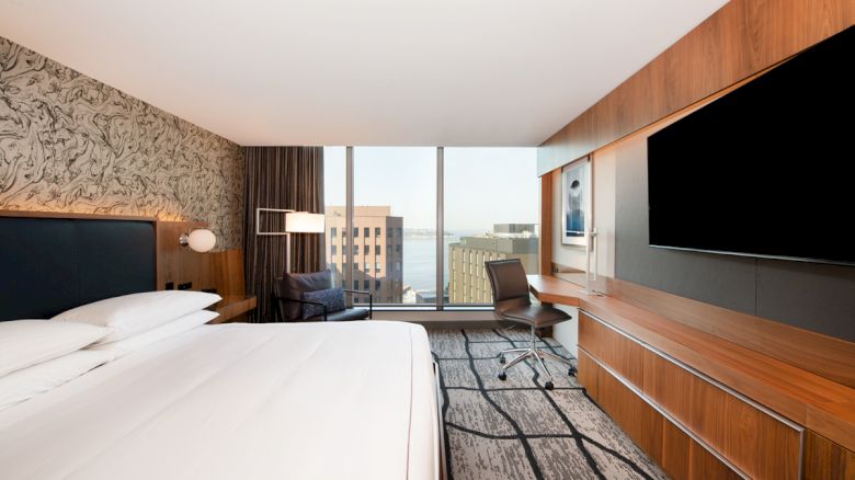 A modern hotel room with a bed, large TV, desk, chair, and a window showing a cityscape view in the background.