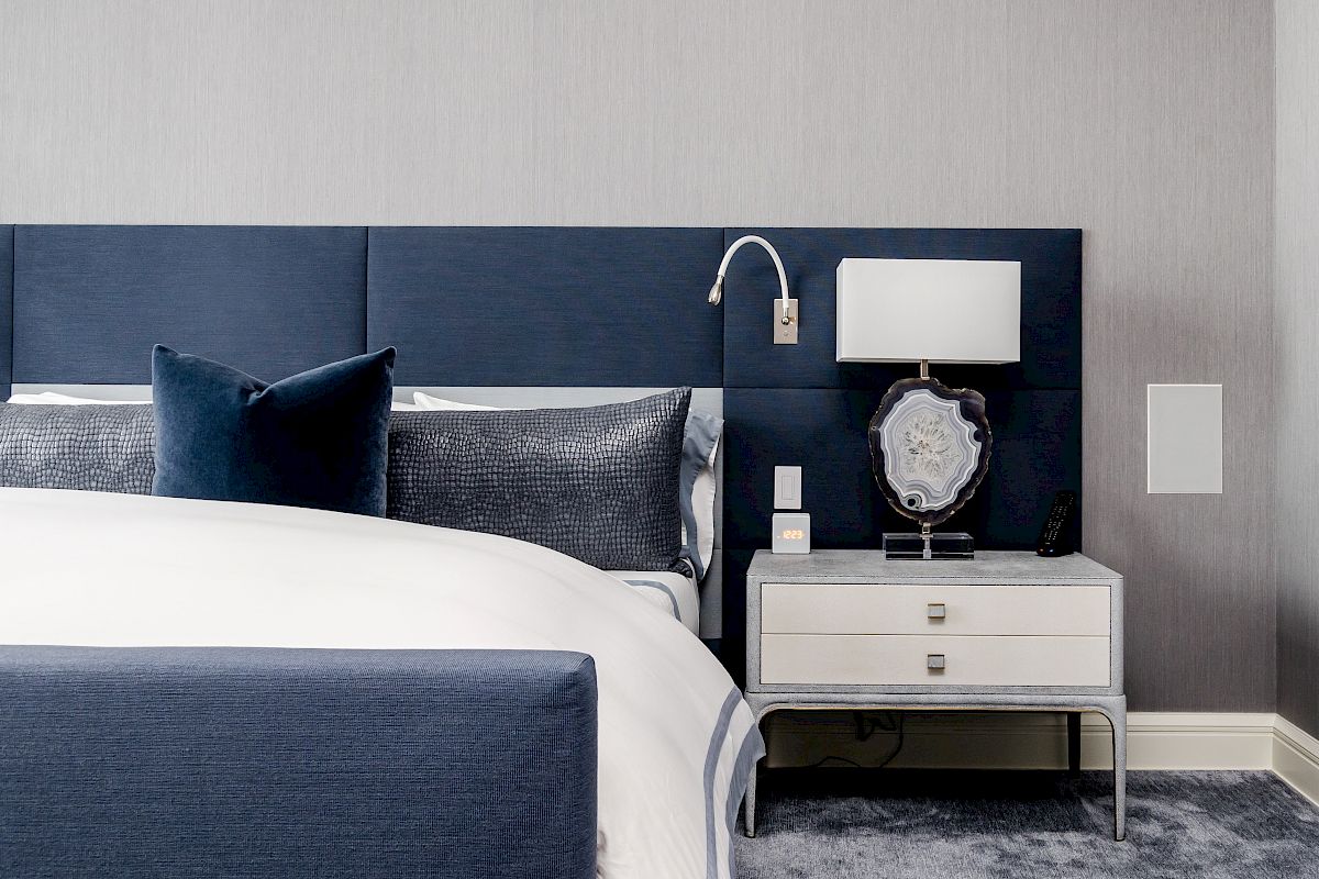 A modern bedroom with a blue headboard, white nightstand, and lamp. A gray and blue color scheme creates a cozy atmosphere.