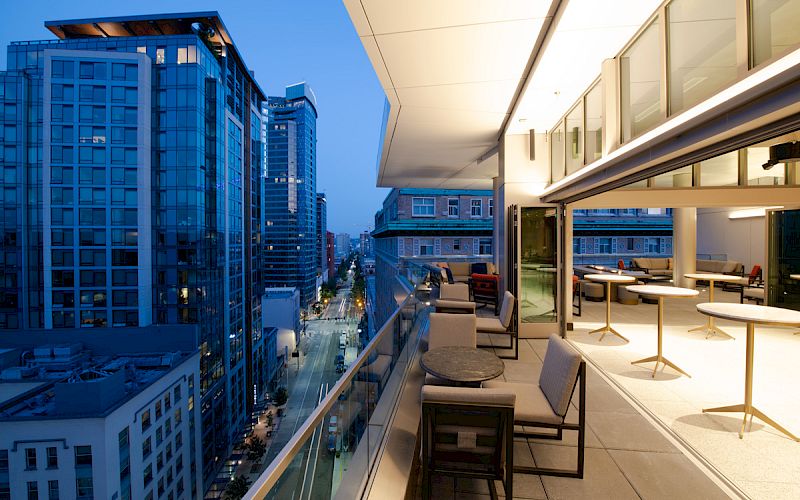 A modern rooftop terrace with outdoor seating overlooks a cityscape at dusk, showcasing tall buildings and a well-lit street below.