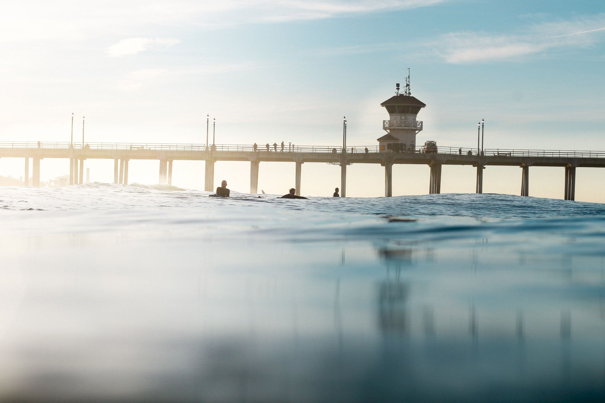 A pier extends over the ocean with people on it, while a few surfers sit in the water in the foreground under a clear sky.